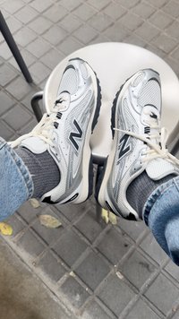 Ténis New Balance brancos e cinzas com atacadores brancos, usados com meias cinzas e jeans azuis, repousando numa banqueta branca sobre pavimento exterior de azulejos.