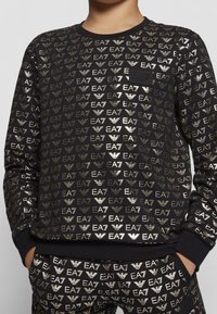 Felpa nera con motivo del logo EA7 in metallo dorato. Presenta polsini e scollo a coste, e sono parzialmente visibili pantaloni coordinati con lo stesso motivo.