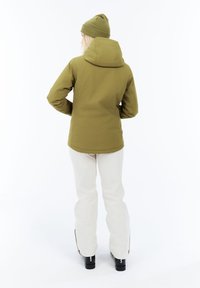 Osoba v olivovo zelenej bundě a klobouku, s bielymi nohavicami a čiernymi čižmami, stojaca chrbtom k fotoaparátu na bielom pozadí.