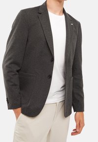 Ciemnoszary blezer wykonany z teksturowanej tkaniny, z dwiema guzikami, klapami i dwiema przednimi kieszeniami, noszony na białym t-shircie.