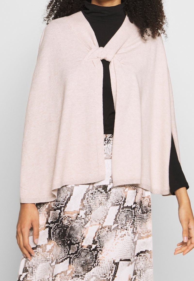 Femme portant une cape beige clair attachée à la poitrine, un haut noir à manches longues et une jupe imprimée serpent.