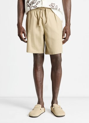 Homme portant un short beige à taille élastique, des chaussures beiges sans lacets, et une chemise blanche avec une broderie florale marron, debout devant un fond blanc.