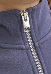 Fialová bunda na zips s kovovým zipsom, na ktorom je vyrazené "JACK & JONES". Tkanina je textúrovaná, s rebrovaným golierom a švami.