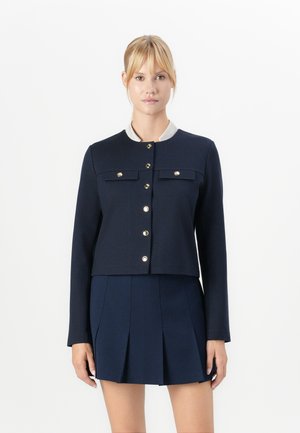 Blonde woman wearing a navy blue long-sleeve buttoned jacket and matching pleated skirt, standing against a white background.