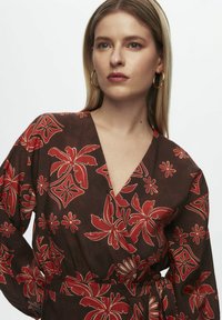 Ruskea kukallinen kietaisumekko, jossa on punaisia trooppisia lehti kuvioita, V-pääntie, pitkät hihat ja sidottu vyötärö. Pehmeä, kevyt kangas.