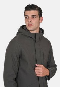 Uomo con capelli corti e scuri che indossa una giacca invernale con cappuccio grigio scuro, che tiene la zona della cerniera frontale, guardando di lato su uno sfondo bianco.