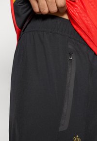 Čierne športové šortky s elastickým pásom, bočnou zipsovou vreckom a zlatým logom blízko lemu. Hladká textura, dĺžka po stred stehna.