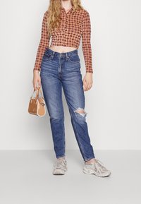 Brązowy wzorzysty krótki top z długim rękawem w połączeniu z niebieskimi jeansami z wysokim stanem z wycięciem na kolanie, dopełnione małą beżową torebką i szarymi trampkami.