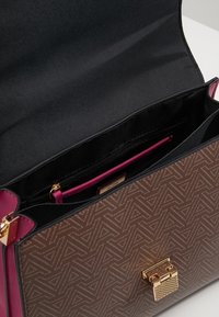 Brązowa torebka w wzory z złotym zapięciem, otwarta, aby pokazać czarne wnętrze z zamykaną kieszenią z różowym wykończeniem.