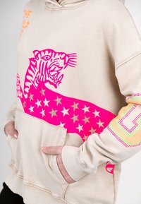 Beige hoodie featuring a pink tiger graphic and a pink star-patterned band. Pocket at the front with ribbed cuffs and a drawstring hood.