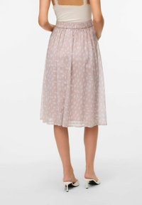 Una donna in piedi, vista da dietro, indossa una gonna rosa chiaro con un motivo di foglie e scarpe con tacco bianche.