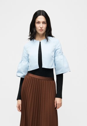 Mujer con el cabello lacio y oscuro, que lleva una chaqueta corta azul claro con mangas abullonadas sobre una blusa negra y una falda plisada marrón.