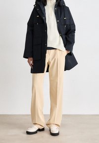 Námořnická parka s více předními kapsami, v kombinaci s krémovým pleteným svetr a béžovými širokými kalhotami. Bílé mokasíny s černými podrážkami.