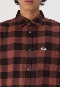 Camisa de franela a cuadros rojos y negros, con botones en la parte delantera, cuello, un bolsillo en el pecho y una pequeña etiqueta blanca de "Wrangler". Textura suave.