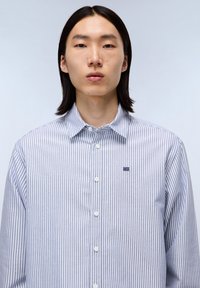 Chemise à rayures bleu clair et blanche, fabriquée en coton. Elle possède un col classique, des boutons sur le devant et un petit logo brodé.
