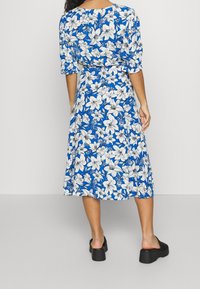 Modré květinové midi šaty s nafouklými rukávy, zdobené bílými a černými květinovými vzory. Spárovány s černými platformovými sandály. Hladká textura.