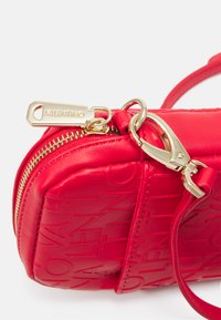 Bolsa de ombro cruzada em pele vermelha com ferragens douradas, apresentando um fecho de zíper e marcação em relevo. Textura suave e design compacto.