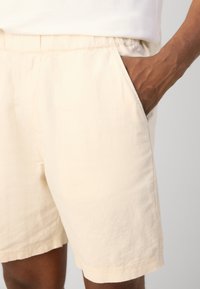 Beige cotton shorts featuring an elastic waistband, side pockets, and a smooth texture. The length is just above the knee.