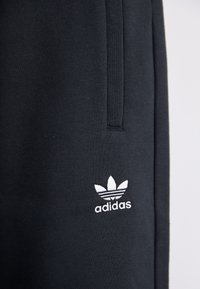 Černé sportovní tepláky vyrobené z texturované tkaniny, s bílým logem Adidas poblíž kapsy. Design zahrnuje boční kapsu.
