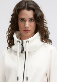 Woman with curly brown hair wears white zip-up hoodie with high collar and drawstrings, facing forward against plain gray background.