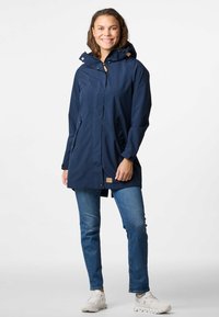 Giacca impermeabile blu scuro con cappuccio, zip frontale, due tasche laterali e una piccola patch con logo sull'orlo, abbinata a jeans blu e sneakers bianche.