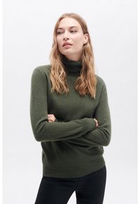 Maglione a collo alto verde oliva in morbido tessuto a maglia. Presenta maniche lunghe e polsini a coste. Indossato con pantaloni neri.