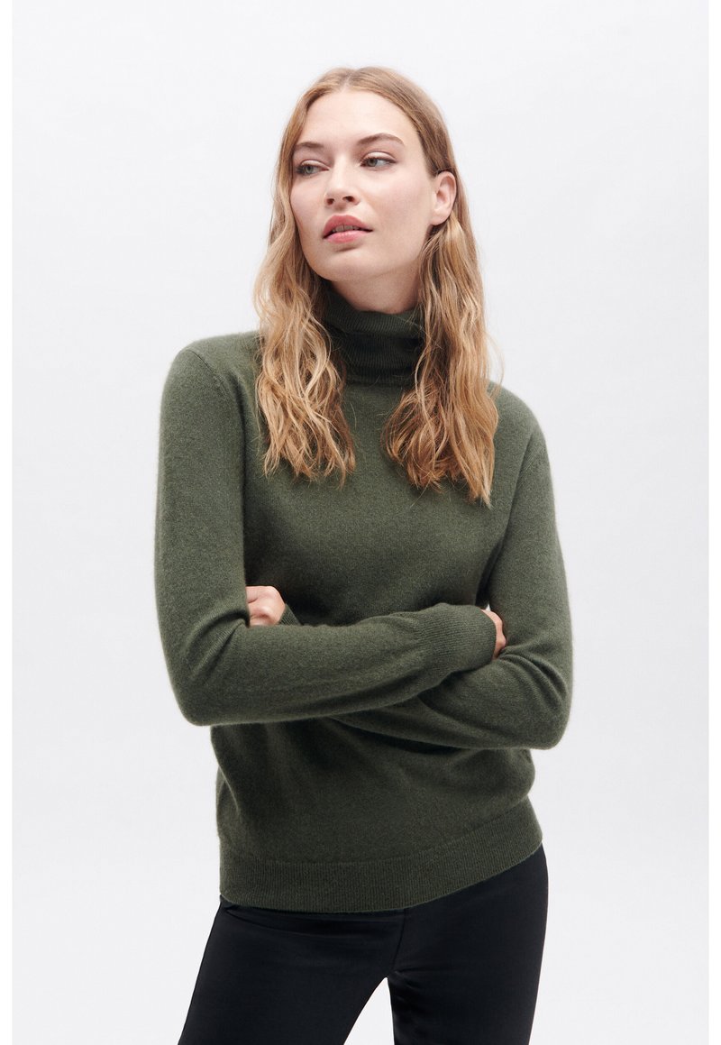 Maglione a collo alto verde oliva in morbido tessuto a maglia. Presenta maniche lunghe e polsini a coste. Indossato con pantaloni neri.