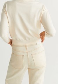 Helles beiges Strickpullover mit einem enganliegenden Saum, kombiniert mit hellbeigen hochgeschnittenen Hosen, die über Gesäßtaschen und kontrastierende Nähte verfügen.