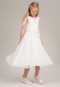 Weißes, plissiertes Kleid mit ärmellosem Design, einem beigen Taillenband und einem fließenden Rock. Kombiniert mit weißen Sneakers mit Spitzenakzenten.