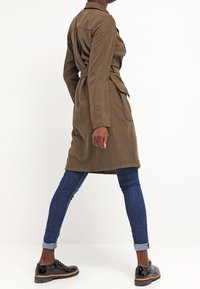Oliwkowy, długi płaszcz trench z długim rękawem wykonany z zamszowego materiału, z talią przewiązaną paskiem. W połączeniu z niebieskimi dżinsami i czarnymi butami na grubej podeszwie.