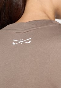 Καφέ αθλητική μπλούζα με υφή υφάσματος, φέρει λευκό λογότυπο Nike και σταυρωμένα γκολφ κλαμπ τυπωμένα στη πλάτη κοντά στον γιακά.