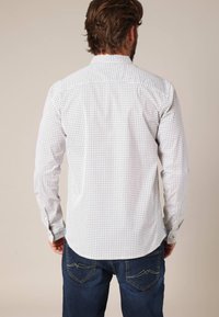 Witte overhemd met een geometrisch patroon, lange mouwen en een puntige kraag. Draagt bij donkerblauwe jeans.