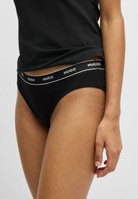 Svart bomulls-bikiniunderkläder med en midjeband som har en logo och vit text. Högskuren design, slät textur och minimalistic stil.