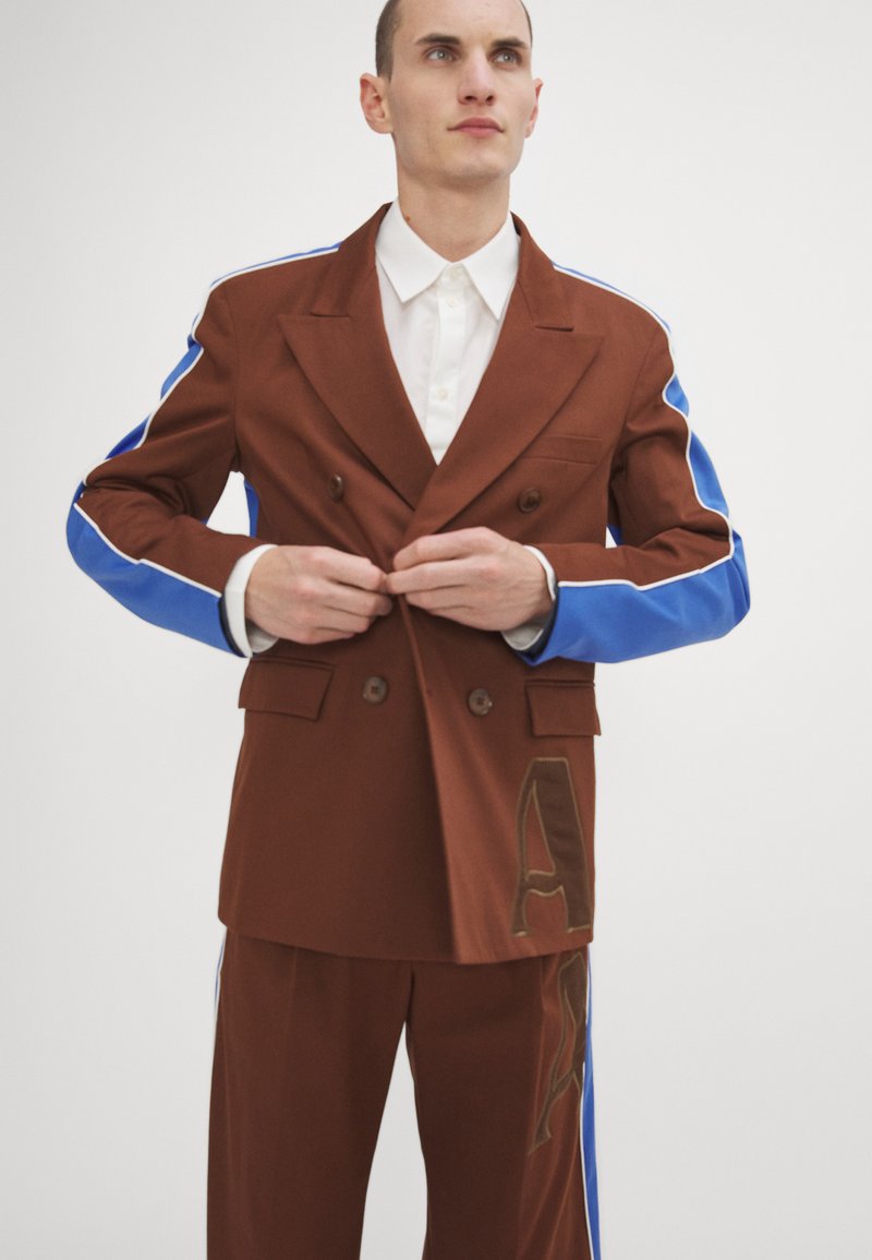 Blazer marrone a doppio petto con accenti blu sulle maniche, abbinato a pantaloni coordinati con un sottile dettaglio del logo. Tessuto liscio e su misura.