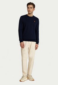 Fiatal férfi áll egy sötétkék, hosszú ujjú pólóban, amelyen egy apró narancssárga logó látható, krém színű nadrágban és bézs sportcipőben, fehér háttér előtt.