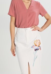 Blush pink kortærmet top med v-hals og knapdetalje, parret med en hvid knælang nederdel med farverig blomsterbroderi og en slid foran.