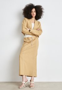 Blazer en lin beige porté sur un pull blanc, associé à une jupe beige assortie avec taille à cordon et détails de couture visibles. Sandales à talons blanches.