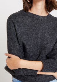 Mörkgrå stickad tröja med rund hals, som visar ett texturerat tyg och lätt uppvikta ärmar. Modellens korsade armar är delvis synliga.