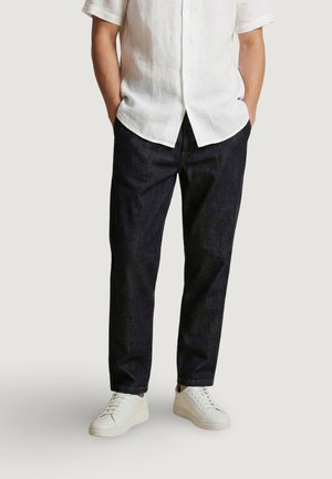 Homme portant une chemise blanche à manches courtes boutonnée, un jean bleu foncé et des baskets blanches, les mains dans les poches sur un fond uni.