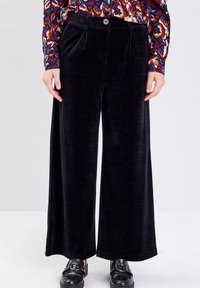Un pantalon large en velours côtelé noir présente une texture lisse, des plis à l'avant et une fermeture à un bouton, associé à des chaussures noires brillantes.