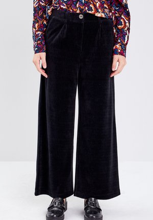 Un pantalon large en velours côtelé noir présente une texture lisse, des plis à l'avant et une fermeture à un bouton, associé à des chaussures noires brillantes.