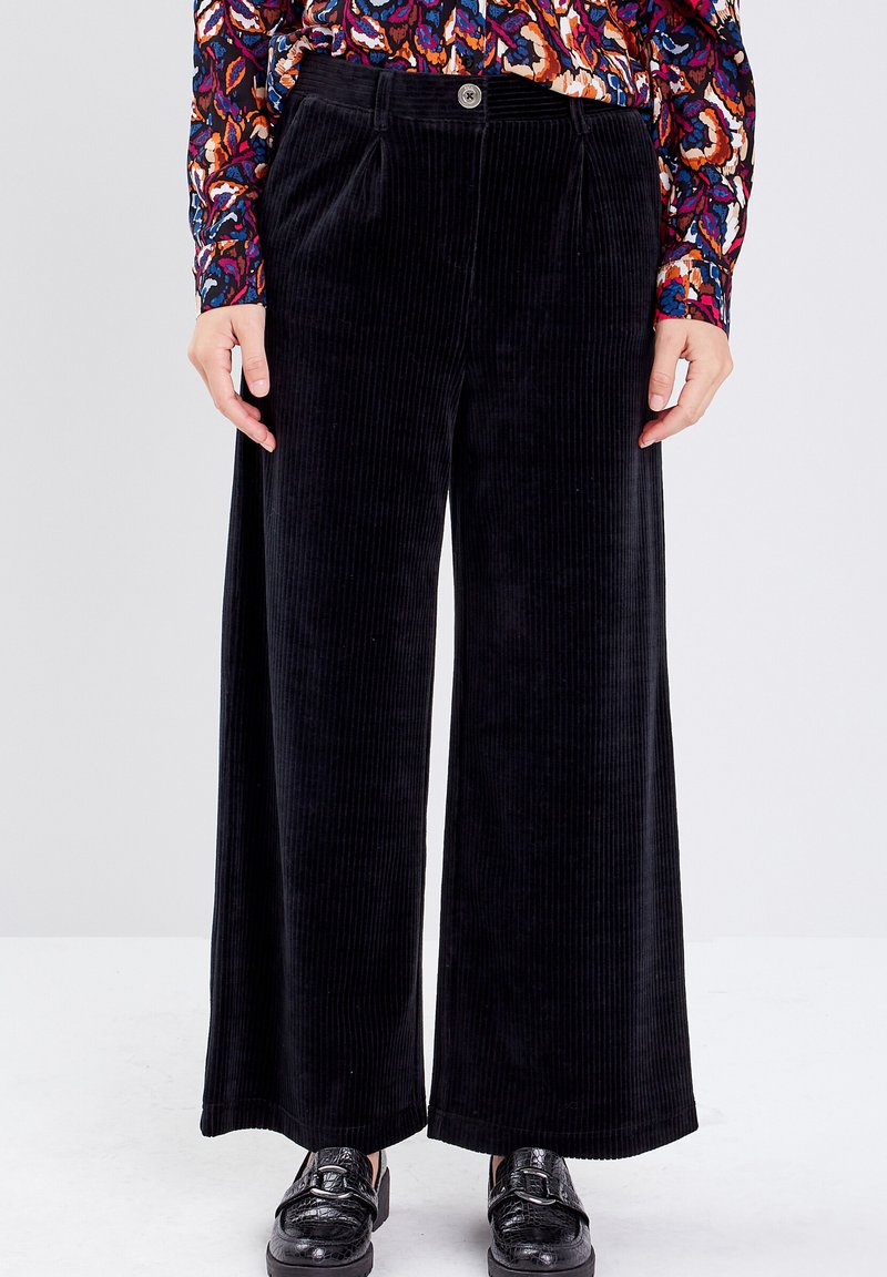 Un pantalon large en velours côtelé noir présente une texture lisse, des plis à l'avant et une fermeture à un bouton, associé à des chaussures noires brillantes.