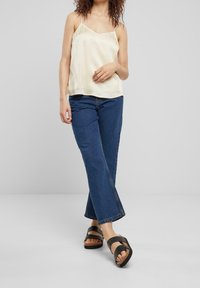 Cream satin camisole with adjustable straps, paired with dark blue flared jeans and black slide sandals featuring a crisscross strap design.