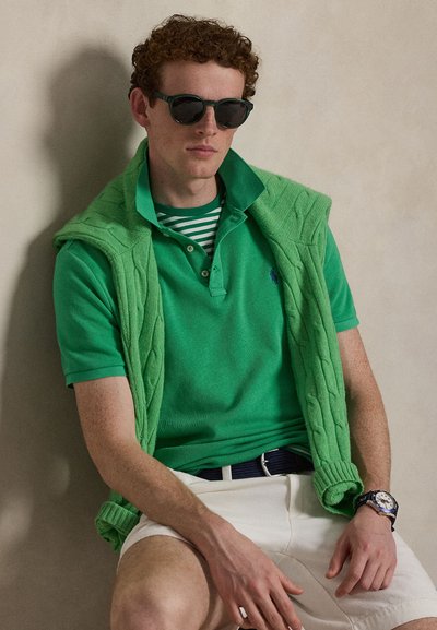 Άνδρας με σγουρά κόκκινα μαλλιά που φοράει μαύρα γυαλιά ηλίου, πράσινα στρωμένα πουκάμισα, κρεμ σορτς και ρολόι χειρός, καθισμένος μπροστά σε μπεζ τοίχο.