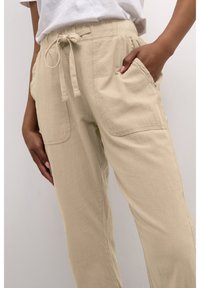 Beige, lockere Hose aus strukturiertem Stoff mit verstellbarem Taillenband, seitlichen Taschen und lässigem Design.