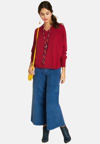 Roter V-Ausschnitt-Pullover, blaue weit geschnittene Hose mit vorderen Taschen, gelbe Handtasche und schwarze Schnürstiefel. Farbenfreudige Perlenkette als Akzent.