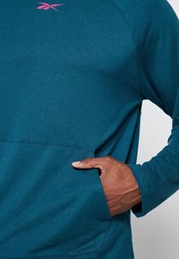 Main dans la poche d'un sweat-shirt bleu sarcelle foncé, mettant en valeur la texture du tissu et le logo rose Reebok sur la poitrine.