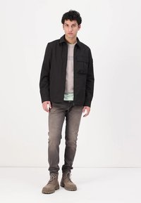 Sort letvægtsjakke med en frontlomme, båret over en grå skjorte og et lyseblåt indre lag. Sammen med grå jeans og beige støvler.