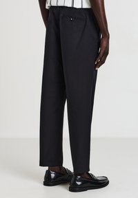 Uomo che indossa pantaloni neri su misura alla caviglia con tasca a bottone sul retro e mocassini in pelle nera, in piedi contro uno sfondo semplice.