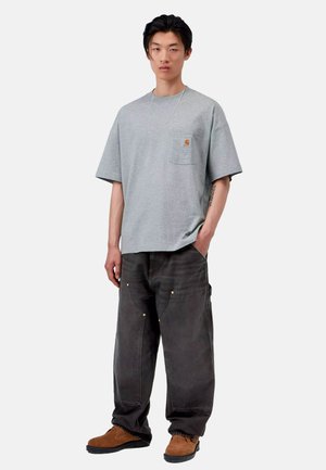 Giovane uomo con capelli neri che indossa una t-shirt grigia oversize, pantaloni neri larghi, scarpe marroni e una collana a catena d'argento, in piedi su sfondo bianco.